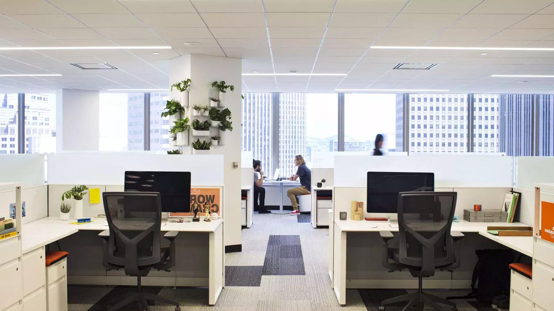 贝博体彩app一座办公大楼的室内场景, 前景有几张桌子，两个人在大窗户前的一张桌子前工作.