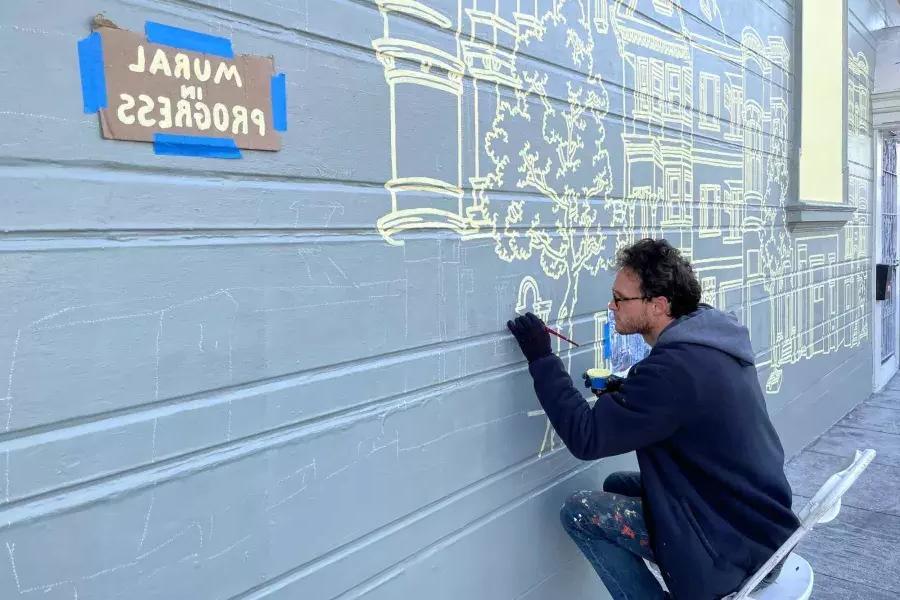 一位艺术家在教会区一栋建筑的侧面画壁画, 在建筑上贴了一个牌子，上面写着“壁画正在进行中”.加州贝博体彩app.
